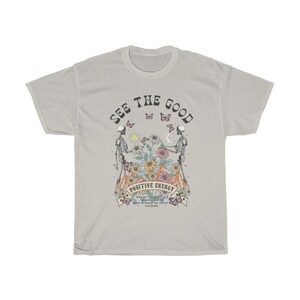 May include: A light grey t-shirt with the words "SEE THE GOOD" arched across the top. The shirt features a graphic of two skeletons holding hands, with flowers and butterflies. The words "POSITIVE ENERGY" are below the graphic.