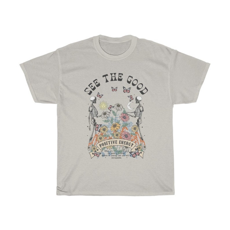 May include: A light grey t-shirt with the words "SEE THE GOOD" arched across the top. The shirt features a graphic of two skeletons holding hands, with flowers and butterflies. The words "POSITIVE ENERGY" are below the graphic.