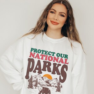 May include: White sweatshirt with a graphic print of a bear in a mountain landscape. The text on the sweatshirt reads "Protect Our National Parks".