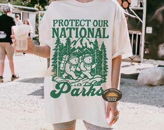 Peut inclure: Un t-shirt blanc avec un motif graphique vert qui dit "Protégez nos parcs nationaux". Le graphique représente deux ours avec des sacs à dos en randonnée dans une forêt. Le texte "Sunset Dreamers Co." et "Original Design" sont également inclus dans le graphique.