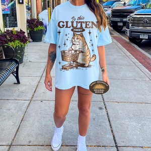May include: Light blue t-shirt with a graphic of a cat pushing a shopping cart, the text "Is this Gluten Free?" is above the cat. The shirt is a relaxed fit.