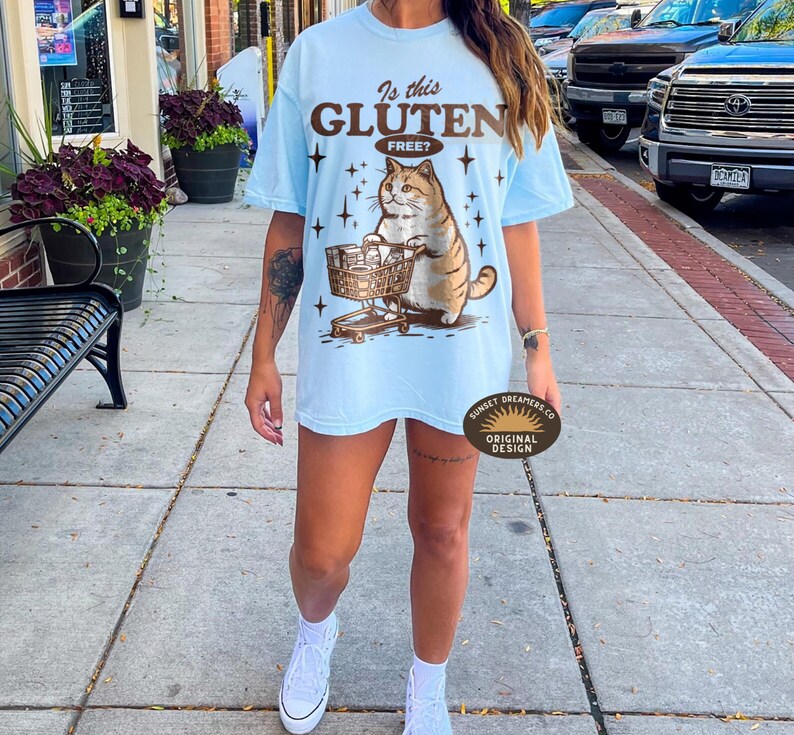 May include: Light blue t-shirt with a graphic of a cat pushing a shopping cart, the text "Is this Gluten Free?" is above the cat. The shirt is a relaxed fit.