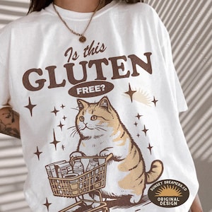 May include: Cream-colored t-shirt with a graphic of a cat pushing a shopping cart filled with groceries. The text "Is this GLUTEN FREE?" is above the cat. The design includes stars and a sun graphic.