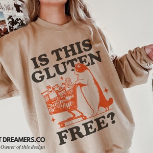 May include: A tan sweatshirt with the text "Is this gluten free?" in black and orange. A cartoon cat is pushing a shopping cart full of groceries.