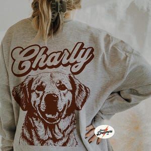 May include: A gray sweatshirt with the word "Charly" in brown script on the back. A brown illustration of a dog's head is below the text.