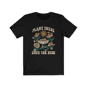 May include: Black t-shirt with a floral design featuring daisies and bees. The text "PLANT THESE SAVE THE BEES" is printed above and below the floral graphic in a vintage style.