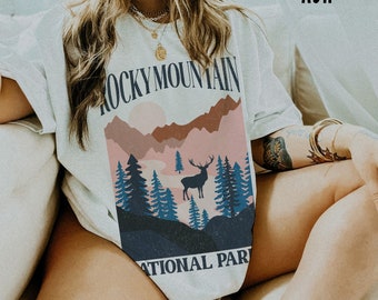 May include: A white t-shirt with a graphic print of a mountain range, a deer, and trees. The text "ROCKY MOUNTAIN NATIONAL PARK" is printed on the shirt.
