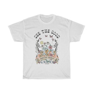 May include: White t-shirt with a graphic design featuring two skeletons, butterflies, and flowers. The text "SEE THE GOOD" is arched across the top, with "POSITIVE ENERGY" below the central image. Additional text is present at the bottom.