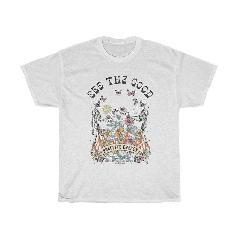 May include: White t-shirt with a graphic design featuring two skeletons, butterflies, and flowers. The text "SEE THE GOOD" is arched across the top, with "POSITIVE ENERGY" below the central image. Additional text is present at the bottom.