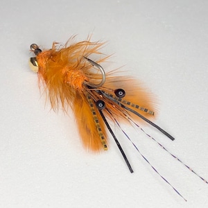 May include: A handmade fishing fly with a hook, featuring orange feathers, black accents, and a gold-colored bead. The fly has a complex design with multiple materials and textures, suitable for fly fishing.