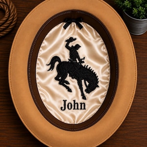 May include: A tan felt hat with a dark brown leather sweatband and a cream-colored satin lining. The interior features a black embroidered graphic of a bucking horse and rider, a small black bow, and the name John embroidered in black thread.