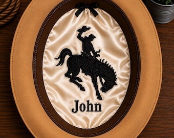 May include: A tan felt hat with a dark brown leather sweatband and a cream-colored satin lining. The interior features a black embroidered graphic of a bucking horse and rider, a small black bow, and the name John embroidered in black thread.