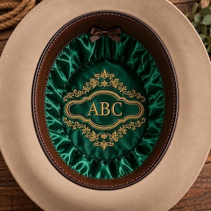 May include: A beige hat with a dark brown leather sweatband and a deep green satin lining. The interior features a gold embroidered filigree frame surrounding the letters ABC in the center, with a small brown bow at the top of the lining.