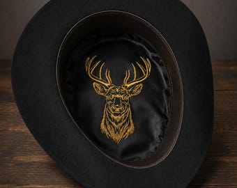 Può includere: Un cappello nero con tesa larga e corona arrotondata. La fodera interna è in raso nero e presenta un dettagliato disegno di testa di cervo ricamato in color oro al centro della corona, circondato da una fascia tergisudore in pelle marrone scuro.