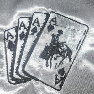 May include: Three playing cards featuring the Ace of Spades, Ace of Diamonds, and Ace of Hearts. The Ace of Hearts card has a black illustration of a cowboy riding a bucking bronco. The cards are embroidered on a silver fabric.
