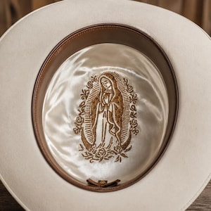 May include: An off-white hat featuring a cream-colored satin lining inside the crown. The lining displays a detailed gold-toned embroidered image of the Virgin of Guadalupe surrounded by roses, with a brown leather sweatband along the inner rim.