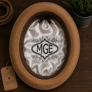 May include: A tan felt hat with a brown leather sweatband and a white satin interior lining. The lining features black embroidered text reading MGE inside an oval frame with decorative leaf accents and a small black bow at the top center.