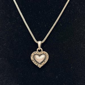 Puede incluir: Collar con colgante de coraz&oacute;n plateado. El colgante en forma de coraz&oacute;n est&aacute; incrustado con peque&ntilde;as piedras preciosas transparentes en el borde. La cadena del collar tiene un dise&ntilde;o delicado y tejido. El colgante cuelga sobre un fondo negro.