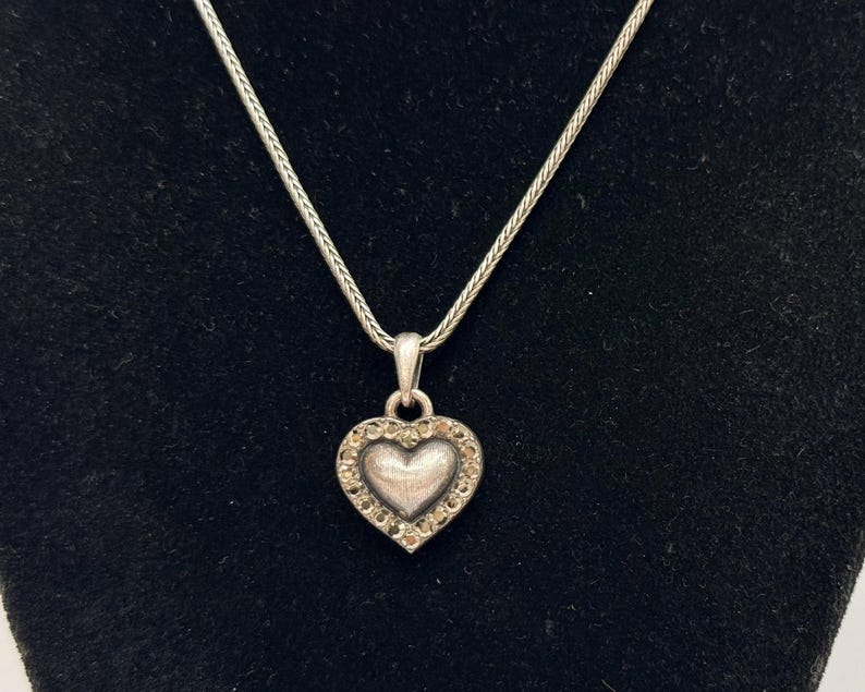 Puede incluir: Collar con colgante de coraz&oacute;n plateado. El colgante en forma de coraz&oacute;n est&aacute; incrustado con peque&ntilde;as piedras preciosas transparentes en el borde. La cadena del collar tiene un dise&ntilde;o delicado y tejido. El colgante cuelga sobre un fondo negro.