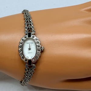 Può includere: Un orologio da polso argentato con un quadrante ovale bianco e un bracciale abbinato. Sul quadrante dell'orologio sono impresse le parole "Majestron" e "Quartz". Il bracciale e la cassa dell'orologio sono adornati con piccole pietre scure.