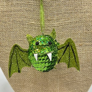 May include: A green bat ornament with glittery wings and a body covered in sequins. The bat has two white fangs and black eyes. A green ribbon is attached for hanging. The ornament is displayed against a burlap background.