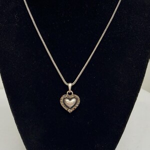 Puede incluir: Un collar plateado con un colgante en forma de coraz&oacute;n. El colgante presenta un coraz&oacute;n central rodeado de peque&ntilde;as piedras transparentes. El collar se muestra sobre un fondo negro.