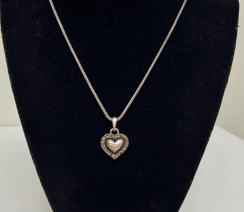 Puede incluir: Un collar plateado con un colgante en forma de coraz&oacute;n. El colgante presenta un coraz&oacute;n central rodeado de peque&ntilde;as piedras transparentes. El collar se muestra sobre un fondo negro.