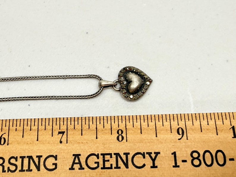Puede incluir: Collar con colgante de coraz&oacute;n plateado. El colgante de coraz&oacute;n est&aacute; incrustado con peque&ntilde;as piedras transparentes alrededor del borde y tiene un centro texturizado. La cadena es de estilo fino y tejido. El colgante mide aproximadamente 2 cm de ancho.