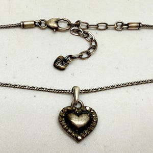 Puede incluir: Collar plateado con colgante en forma de coraz&oacute;n. El colgante presenta un coraz&oacute;n texturizado en el centro y un borde de peque&ntilde;os detalles brillantes. La cadena tiene un dise&ntilde;o delicado y tejido, e incluye una cadena ajustable.