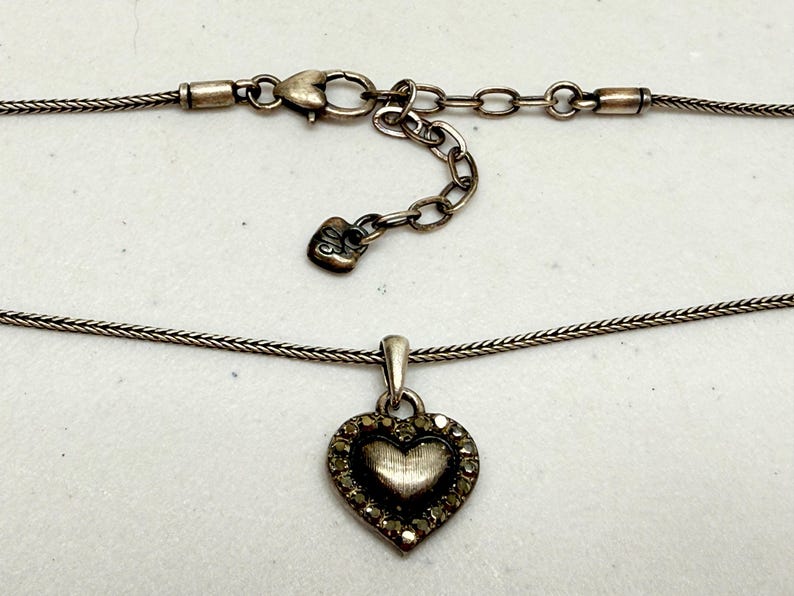 Puede incluir: Collar plateado con colgante en forma de coraz&oacute;n. El colgante presenta un coraz&oacute;n texturizado en el centro y un borde de peque&ntilde;os detalles brillantes. La cadena tiene un dise&ntilde;o delicado y tejido, e incluye una cadena ajustable.
