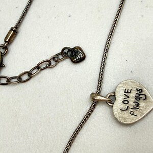 Puede incluir: Collar plateado con colgante en forma de coraz&oacute;n. El colgante lleva grabadas las palabras "Love Always" en negro. El collar tiene una cadena ajustable y un cierre de mosquet&oacute;n.