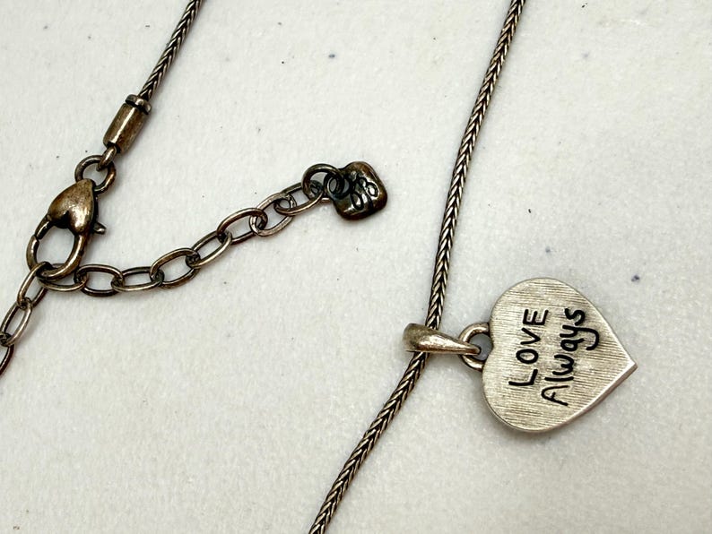 Puede incluir: Collar plateado con colgante en forma de coraz&oacute;n. El colgante lleva grabadas las palabras "Love Always" en negro. El collar tiene una cadena ajustable y un cierre de mosquet&oacute;n.