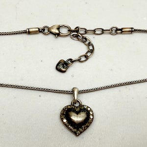 Puede incluir: Collar plateado con colgante en forma de coraz&oacute;n. El colgante est&aacute; adornado con peque&ntilde;as piedras transparentes en el borde. La cadena tiene un fino dise&ntilde;o tejido y se cierra con un cierre de mosquet&oacute;n.