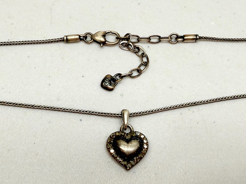 Puede incluir: Collar plateado con colgante en forma de coraz&oacute;n. El colgante est&aacute; adornado con peque&ntilde;as piedras transparentes en el borde. La cadena tiene un fino dise&ntilde;o tejido y se cierra con un cierre de mosquet&oacute;n.