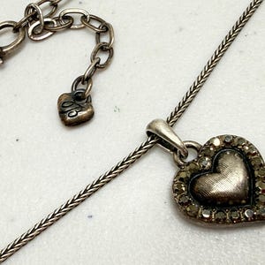 Puede incluir: Collar con colgante de coraz&oacute;n plateado. El colgante en forma de coraz&oacute;n est&aacute; adornado con peque&ntilde;as piedras de color oscuro alrededor del borde. El collar tiene una cadena delicada y un cierre en forma de coraz&oacute;n. Tambi&eacute;n es visible un peque&ntilde;o dije de coraz&oacute;n con una inicial.