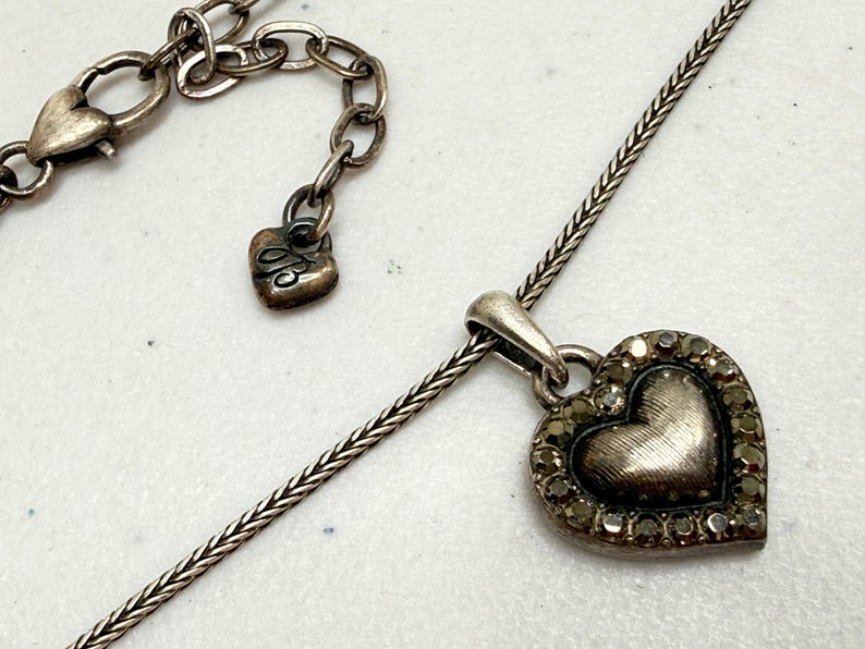 Puede incluir: Collar con colgante de coraz&oacute;n plateado. El colgante en forma de coraz&oacute;n est&aacute; adornado con peque&ntilde;as piedras de color oscuro alrededor del borde. El collar tiene una cadena delicada y un cierre en forma de coraz&oacute;n. Tambi&eacute;n es visible un peque&ntilde;o dije de coraz&oacute;n con una inicial.