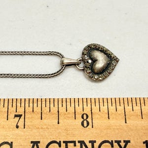 Puede incluir: Un collar plateado con un colgante en forma de coraz&oacute;n. El colgante tiene un coraz&oacute;n texturizado en el centro, rodeado de peque&ntilde;as piedras brillantes. La cadena es de dise&ntilde;o fino y tejido. El collar se muestra sobre una superficie blanca.