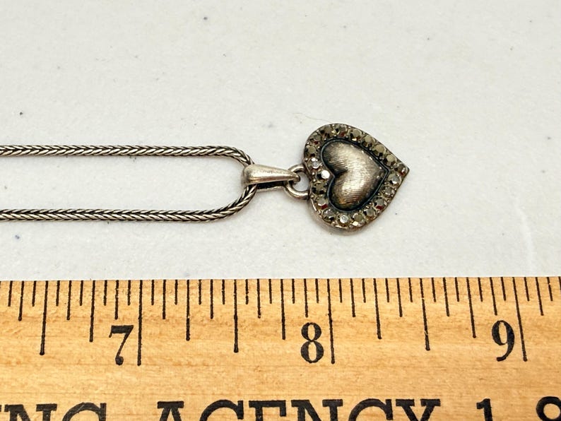 Puede incluir: Un collar plateado con un colgante en forma de coraz&oacute;n. El colgante tiene un coraz&oacute;n texturizado en el centro, rodeado de peque&ntilde;as piedras brillantes. La cadena es de dise&ntilde;o fino y tejido. El collar se muestra sobre una superficie blanca.