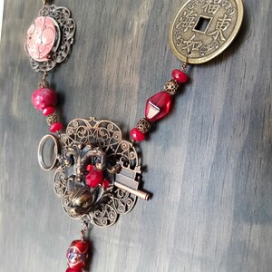 May include: A red and gold necklace with a key, a frog, and a Chinese coin with the characters "福" and "財" on it. The necklace is made of beads, metal, and filigree.