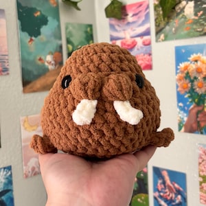 May include: A handmade brown crochet walrus plush toy with white tusks and black eyes. The plush is held in a hand, showcasing its textured, rounded form. The background features art prints and decorative vines.