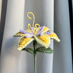 May include: A handmade crochet flower featuring yellow and lilac petals, green leaves, and a yellow centre. The stem is wrapped in green yarn. The flower is set against a white backdrop.