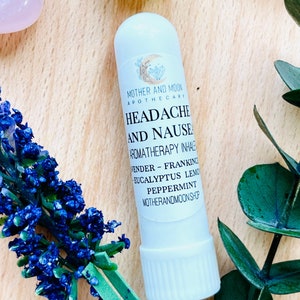 May include: White aromatherapy inhaler tube with a label that reads "MOTHER AND MOON APOTHECARY HEADACHE AND NAUSEA AROMATHERAPY INHALE LAVENDER - FRANKINCENSE EUCALYPTUS LEMON PEPPERMINT MOTHERANDMOONSHOP".