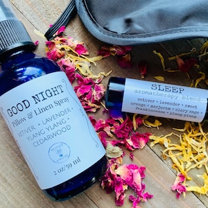 May include: A set of two aromatherapy products for sleep. A blue glass bottle with a white label that reads "Good Night Pillow & Linen Spray" with a list of essential oils: vetiver, lavender, ylang ylang, and cedarwood. A brown glass roller bottle with a white label that reads "Sleep aromatherapy blend" with a list of essential oils: vetiver, lavender, sweet orange, marjoram, clary sage, frankincense, and ylang ylang.