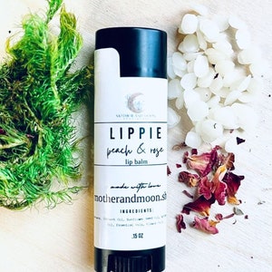 May include: A black and white lip balm tube with the text "LIPPIE peach & rose lip balm" and "motherandmoon.sh" printed on the label. The tube is sitting on a wooden surface with white wax beads and dried rose petals.