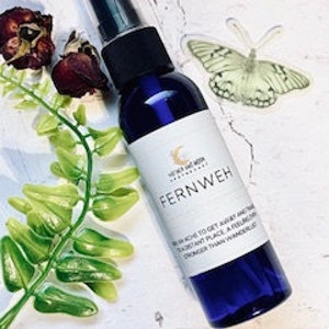 May include: A blue glass bottle with a spray nozzle, labeled "FERNWEH" and containing a clear liquid. The bottle is surrounded by green leaves and a dried rose.