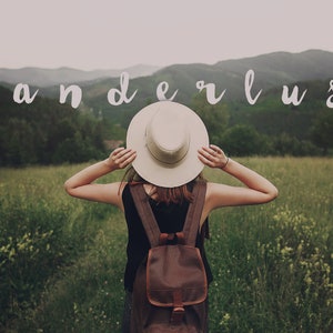 May include: A person wearing a white hat and a brown backpack stands in a field with green grass and mountains in the background. The word "wanderlust" is written in white letters across the top of the image.
