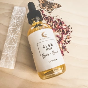 Peut inclure: Un flacon en verre transparent avec une &eacute;tiquette blanche sur laquelle est inscrit "Glow Serum - Rosehip - Rosemar" et "2 fl oz / 50 ml". Le flacon est pos&eacute; sur une surface en bois brun clair avec un cristal blanc et des p&eacute;tales de rose s&eacute;ch&eacute;s.