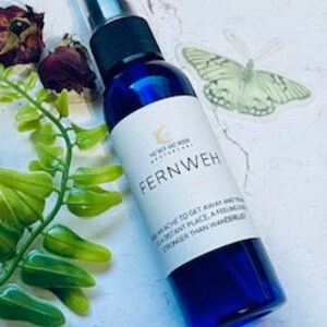 May include: A blue glass bottle with a white label that reads "Fernweh" and a description of the word. The bottle has a black spray nozzle and is sitting on a white surface with green leaves and a butterfly sticker.