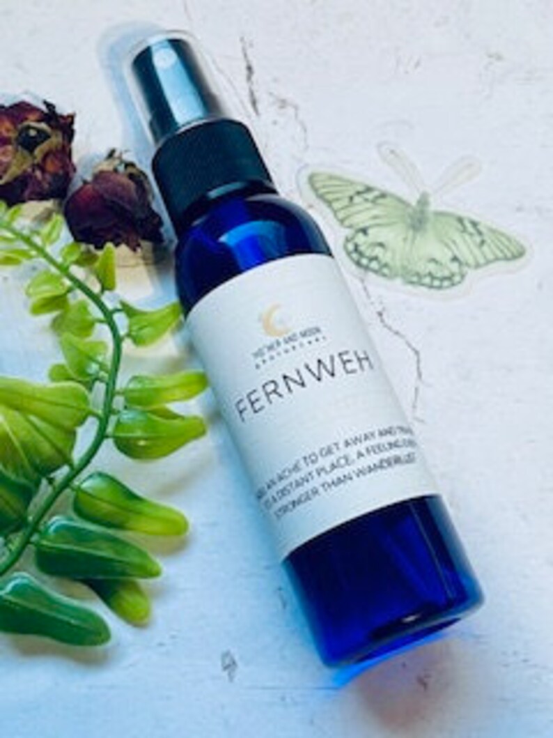May include: A blue glass bottle with a white label that reads "Fernweh" and a description of the word. The bottle has a black spray nozzle and is sitting on a white surface with green leaves and a butterfly sticker.