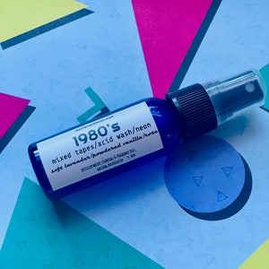 May include: A blue glass bottle with a black spray nozzle and white text that reads "1980's mixed tapes/acid wash/neon soft lavender/powdered vanilla/rose" and "DISTILLED WATER, ESSENTIAL & FRAGRANCE OILS, NATURAL PRESERVATIVE 1 OZ".